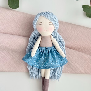 May include: A handmade doll with light blue yarn hair and a smiling face. The doll wears a tan top and a blue skirt with a white floral pattern. It has light brown boots and white arms and legs. The doll is a soft toy.
