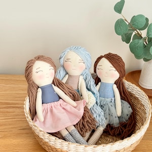May include: Three handmade cloth dolls with yarn hair sit in a woven basket. The dolls have smiling faces and wear dresses in shades of blue and pink. One doll has a blue tulle skirt with star accents. The basket is made of natural fibers.