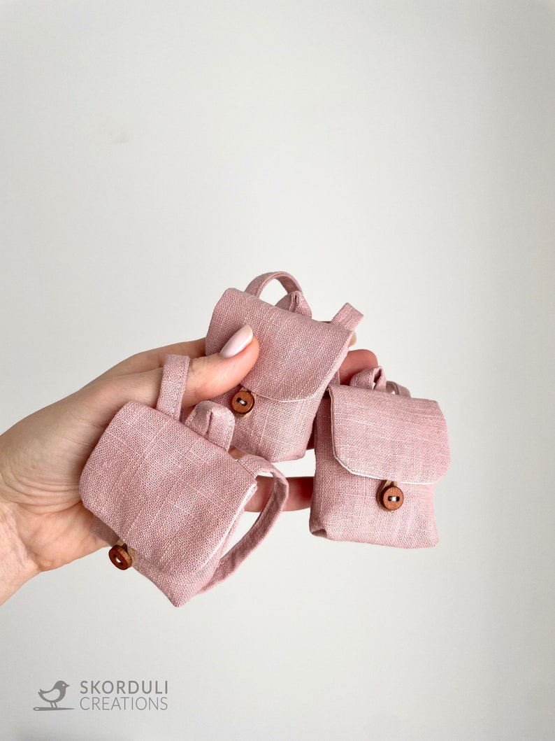 May include: Three miniature pink linen backpacks with brown wooden buttons. The backpacks are arranged in a hand, with the largest backpack in the center and the two smaller backpacks on either side.