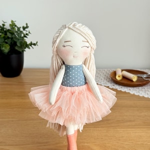 May include: A handmade doll with a light complexion, blonde yarn hair, and a smiling face. The doll wears a blue polka-dot top, a pink tulle skirt, and pink boots. The doll is a charming, handcrafted item.