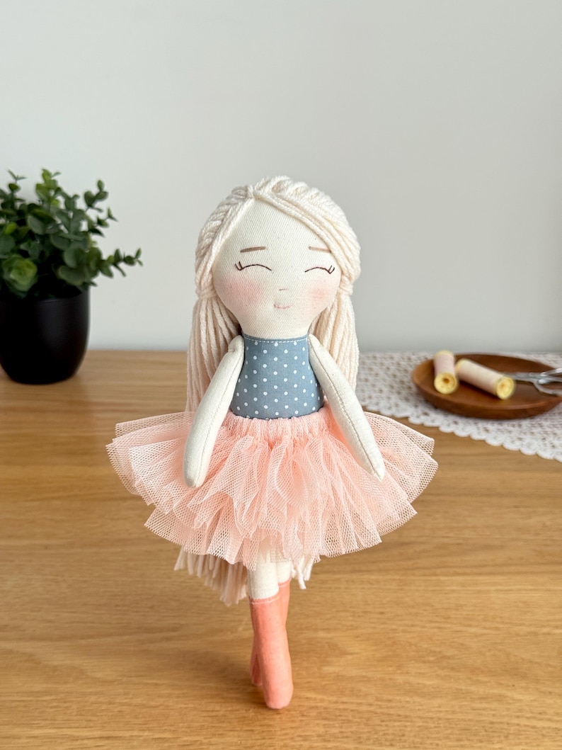 May include: A handmade doll with a light complexion, blonde yarn hair, and a smiling face. The doll wears a blue polka-dot top, a pink tulle skirt, and pink boots. The doll is a charming, handcrafted item.