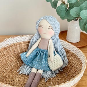 May include: A handmade doll with light blue yarn hair, rosy cheeks, and a blue patterned skirt. The doll is holding a small, white crocheted bag and is seated in a woven basket with a white painted rim. The doll's socks are brown.