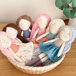 May include: A collection of handmade fabric dolls with yarn hair in a woven basket. The dolls have various hair colors, including pink, brown, and blonde. They wear dresses in shades of blue, pink, and white. The dolls are arranged in a basket on a wooden surface.