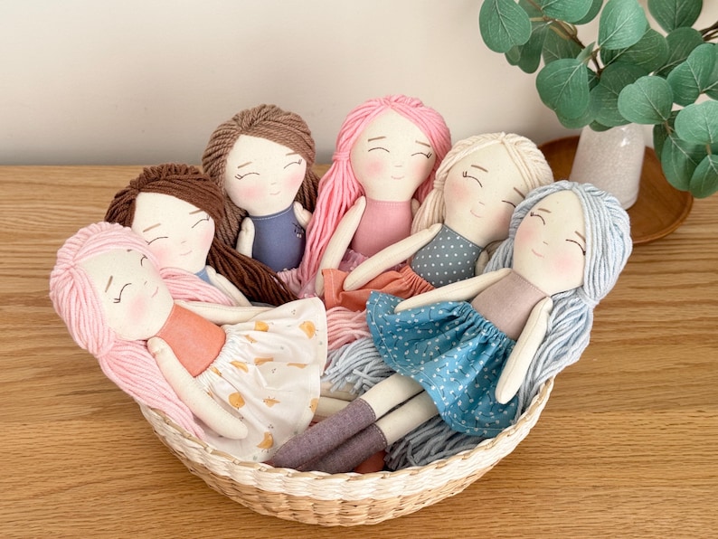 May include: A collection of handmade fabric dolls with yarn hair in a woven basket. The dolls have various hair colors, including pink, brown, and blonde. They wear dresses in shades of blue, pink, and white. The dolls are arranged in a basket on a wooden surface.