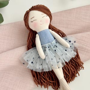 May include: A handmade doll with brown yarn hair and a blue bodice. The doll wears a light blue tulle skirt with dark blue star patterns. The doll's legs are beige with striped socks. The doll is laying on a pink fabric.