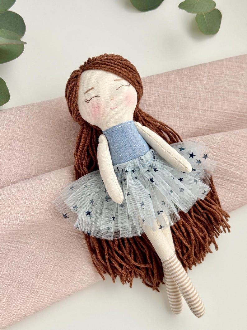 May include: A handmade doll with brown yarn hair and a blue bodice. The doll wears a light blue tulle skirt with dark blue star patterns. The doll's legs are beige with striped socks. The doll is laying on a pink fabric.