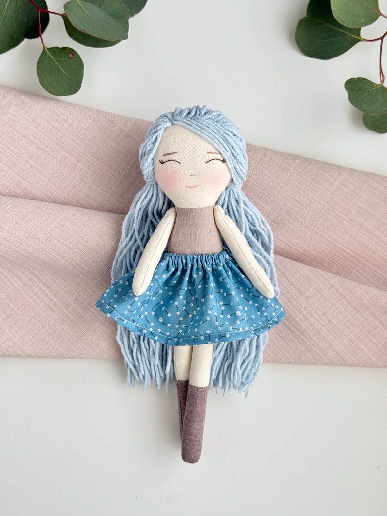 May include: A handmade doll with light blue yarn hair and a smiling face. The doll wears a tan top and a blue skirt with a white floral pattern. It has light brown boots and white arms and legs. The doll is a soft toy.