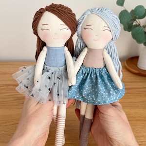 May include: Two handmade cloth dolls with smiling faces. One doll has brown yarn hair, a blue dress, and a tulle skirt with star accents. The other doll has blue yarn hair and a blue floral skirt. Both dolls have striped socks.