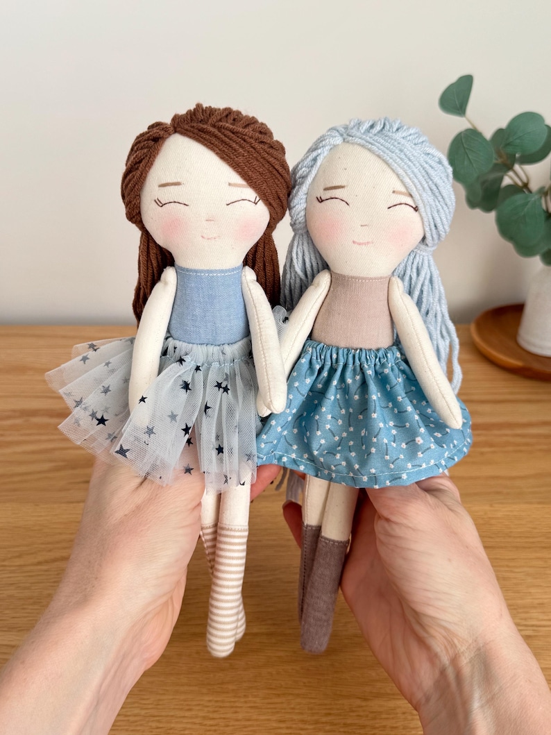 May include: Two handmade cloth dolls with smiling faces. One doll has brown yarn hair, a blue dress, and a tulle skirt with star accents. The other doll has blue yarn hair and a blue floral skirt. Both dolls have striped socks.