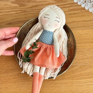 May include: A handmade doll with long, light blonde yarn hair, rosy cheeks, and closed eyes. The doll wears a blue polka-dot top and a peach-colored skirt and socks. A sprig of greenery is tucked into the doll's bodice. The doll is in a decorative metal bowl.