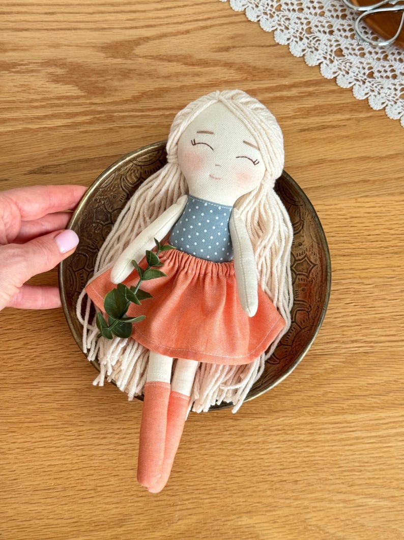 May include: A handmade doll with long, light blonde yarn hair, rosy cheeks, and closed eyes. The doll wears a blue polka-dot top and a peach-colored skirt and socks. A sprig of greenery is tucked into the doll's bodice. The doll is in a decorative metal bowl.