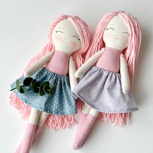 K&ouml;nnte beinhalten: Zwei handgefertigte Stoffpuppen mit rosafarbenem Garnhaar, rosa-wei&szlig;en Kleidern und rosa Socken. Die Puppen sitzen auf einer wei&szlig;en Oberfl&auml;che.