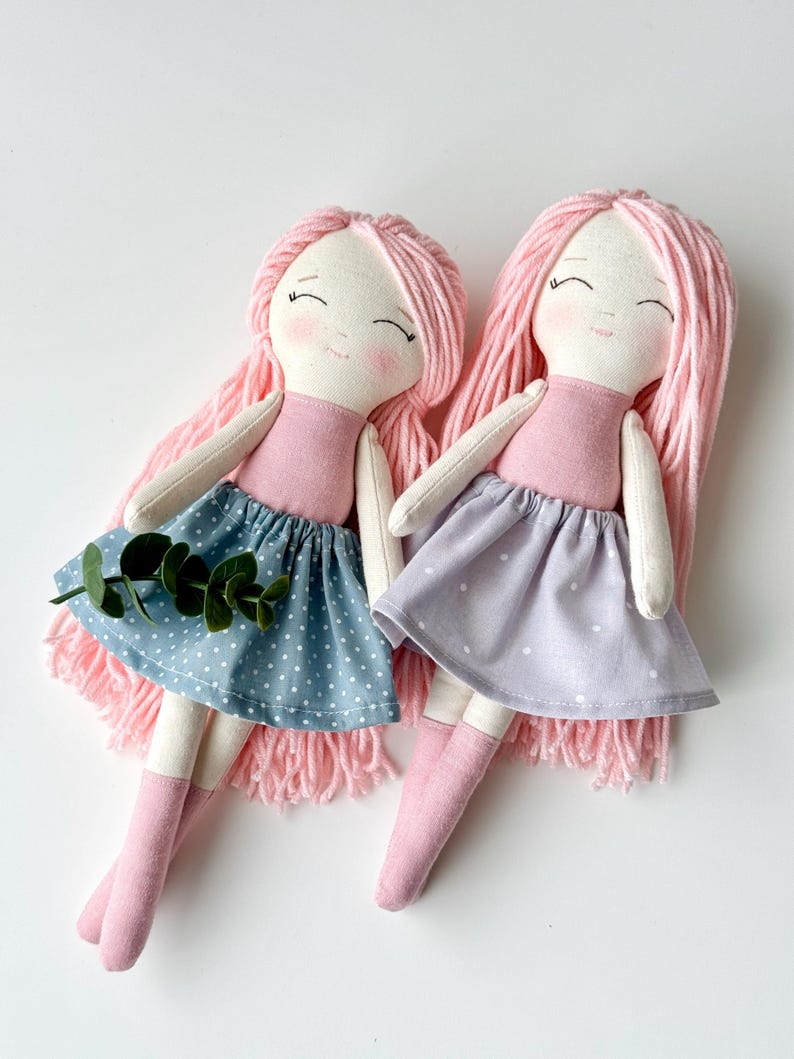 K&ouml;nnte beinhalten: Zwei handgefertigte Stoffpuppen mit rosafarbenem Garnhaar, rosa-wei&szlig;en Kleidern und rosa Socken. Die Puppen sitzen auf einer wei&szlig;en Oberfl&auml;che.