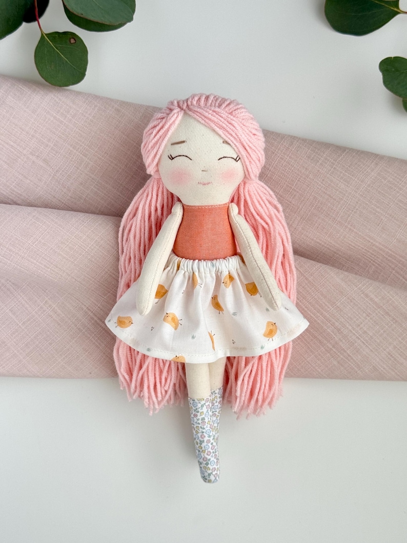 May include: A handmade cloth doll with pink yarn hair and a peach-colored top. The doll wears a white skirt with a yellow bird pattern and floral print boots. The doll has a sweet, smiling face.