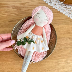 May include: Handmade cloth doll with pink yarn hair, a peach-colored top, and a white skirt with a yellow pattern. The doll has rosy cheeks and closed eyes. It is placed in a decorative brass bowl. The doll's legs are covered with blue floral fabric.
