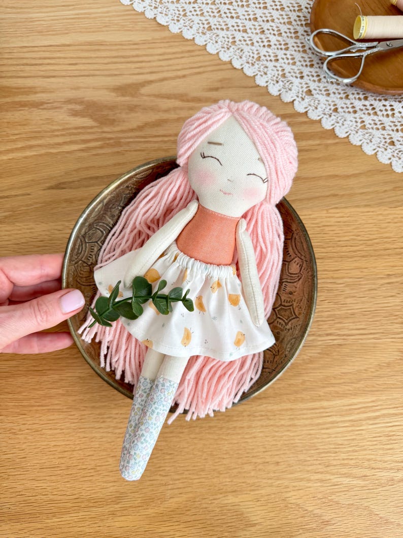 May include: Handmade cloth doll with pink yarn hair, a peach-colored top, and a white skirt with a yellow pattern. The doll has rosy cheeks and closed eyes. It is placed in a decorative brass bowl. The doll's legs are covered with blue floral fabric.