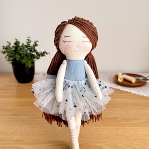 May include: A handmade doll with brown yarn hair and a blue bodice. The doll wears a white tulle skirt with blue star accents and striped socks. The doll has a sweet, smiling face and is a charming decorative item.