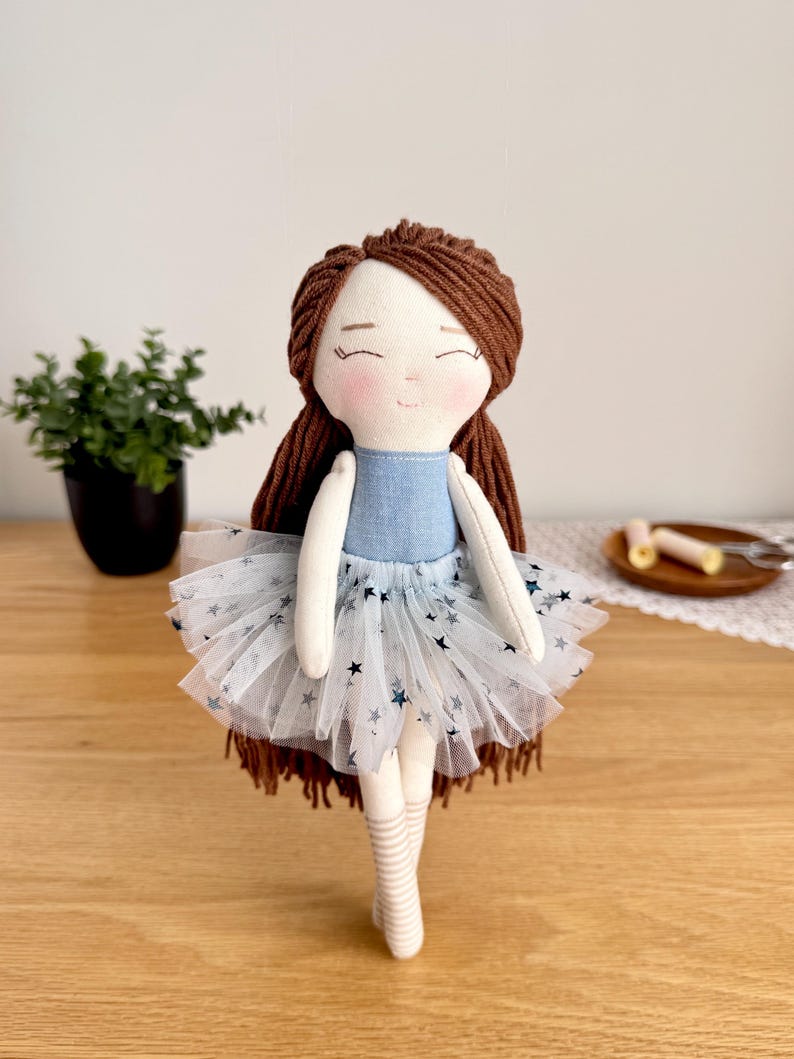 May include: A handmade doll with brown yarn hair and a blue bodice. The doll wears a white tulle skirt with blue star accents and striped socks. The doll has a sweet, smiling face and is a charming decorative item.