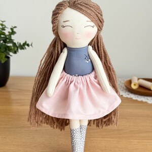 Peut inclure: Poup&eacute;e en tissu faite &agrave; la main avec des cheveux en fil marron et un visage souriant. La poup&eacute;e porte un haut bleu, une jupe rose et des bottes &agrave; motifs gris et blancs. C'est un jouet doux.