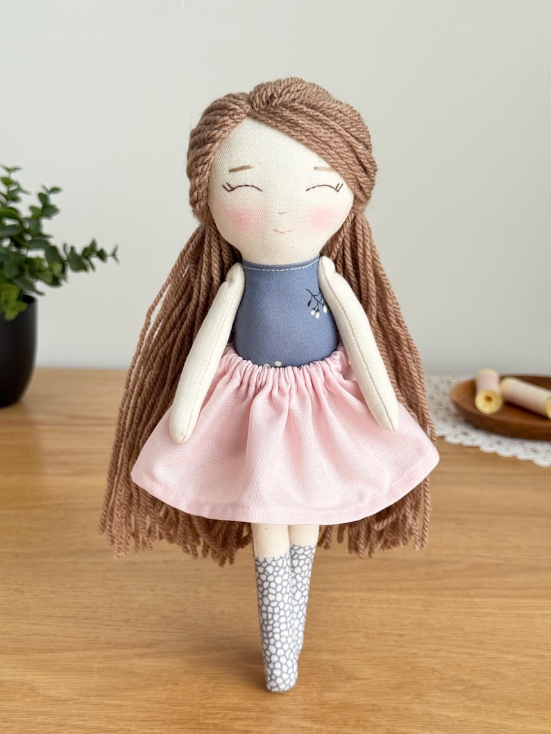 Peut inclure: Poup&eacute;e en tissu faite &agrave; la main avec des cheveux en fil marron et un visage souriant. La poup&eacute;e porte un haut bleu, une jupe rose et des bottes &agrave; motifs gris et blancs. C'est un jouet doux.