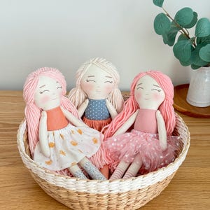 May include: Three handmade cloth dolls with yarn hair, each in a different outfit, sit in a woven basket. One doll has pink hair and a peach top with a white skirt. Another has blonde hair and a blue polka dot top. The third doll has pink hair and a pink tutu.