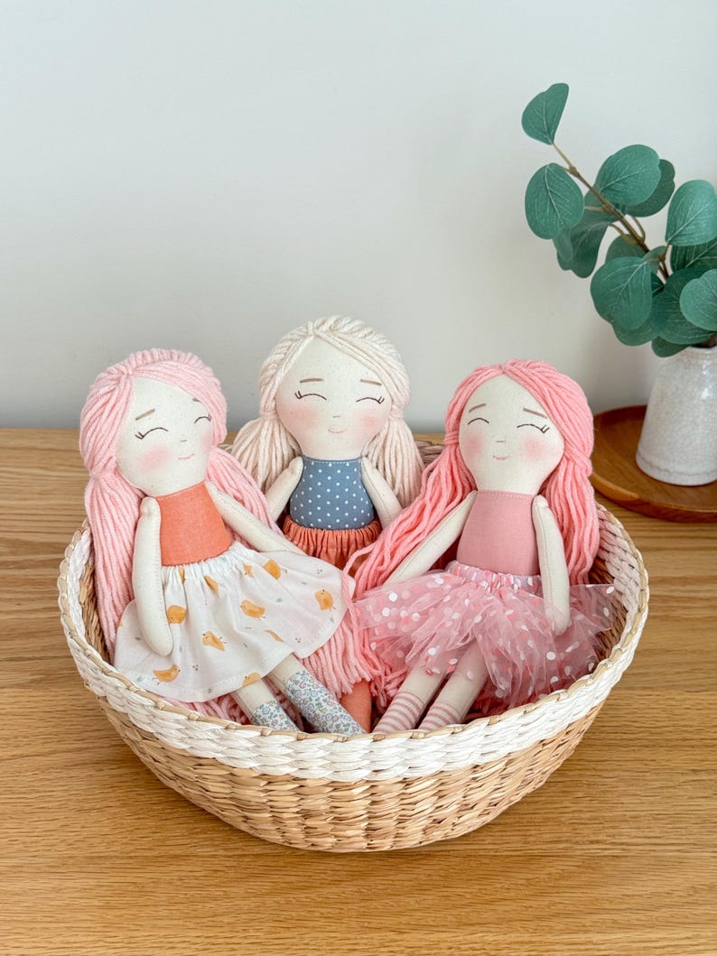 May include: Three handmade cloth dolls with yarn hair, each in a different outfit, sit in a woven basket. One doll has pink hair and a peach top with a white skirt. Another has blonde hair and a blue polka dot top. The third doll has pink hair and a pink tutu.