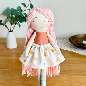 May include: A handmade fabric doll with pink yarn hair and a peach-colored top. The doll wears a white skirt with orange bird illustrations and floral patterned boots. The doll has a smiling face with closed eyes. The doll is a children's toy.