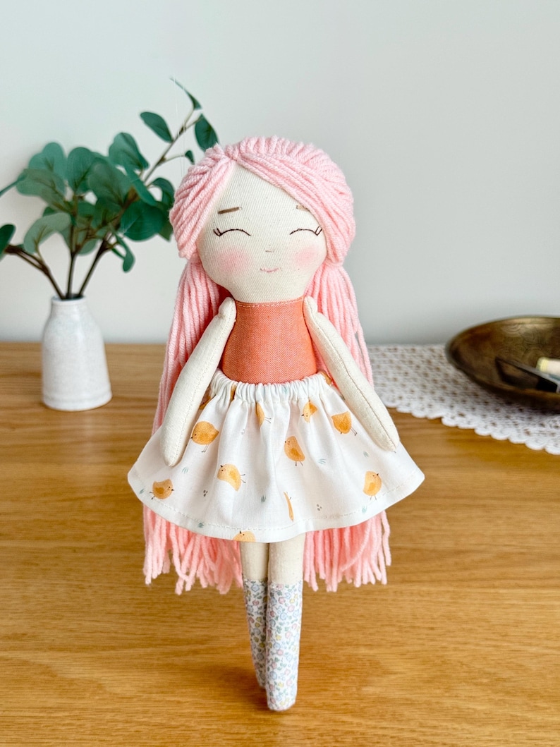 May include: A handmade fabric doll with pink yarn hair and a peach-colored top. The doll wears a white skirt with orange bird illustrations and floral patterned boots. The doll has a smiling face with closed eyes. The doll is a children's toy.