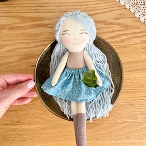 May include: A handmade doll with light blue yarn hair, a tan top, and a blue polka-dot skirt. The doll has a serene expression and is holding a small green plant. It is set in a decorative brass dish.