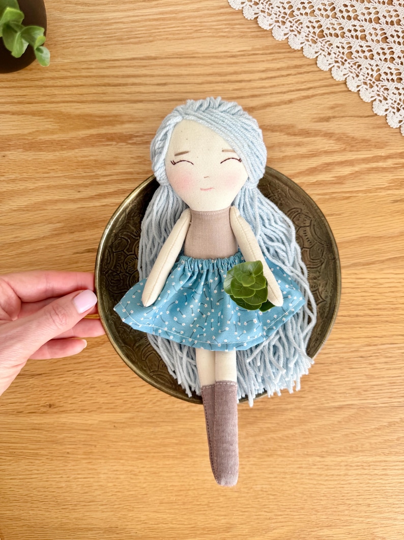 May include: A handmade doll with light blue yarn hair, a tan top, and a blue polka-dot skirt. The doll has a serene expression and is holding a small green plant. It is set in a decorative brass dish.