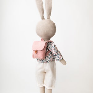 May include: A plush bunny doll wearing a pink backpack, a white shirt with a floral pattern, and white pants. The bunny doll is standing on a wooden base.
