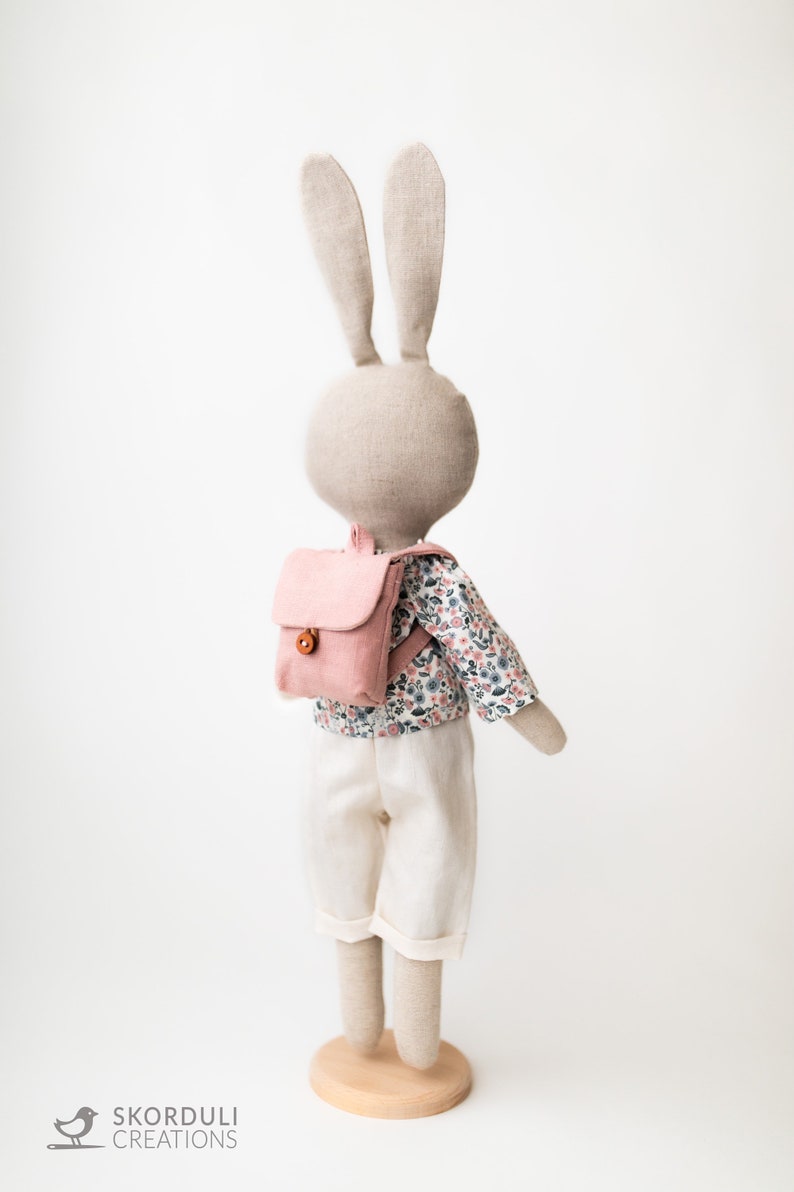 May include: A plush bunny doll wearing a pink backpack, a white shirt with a floral pattern, and white pants. The bunny doll is standing on a wooden base.