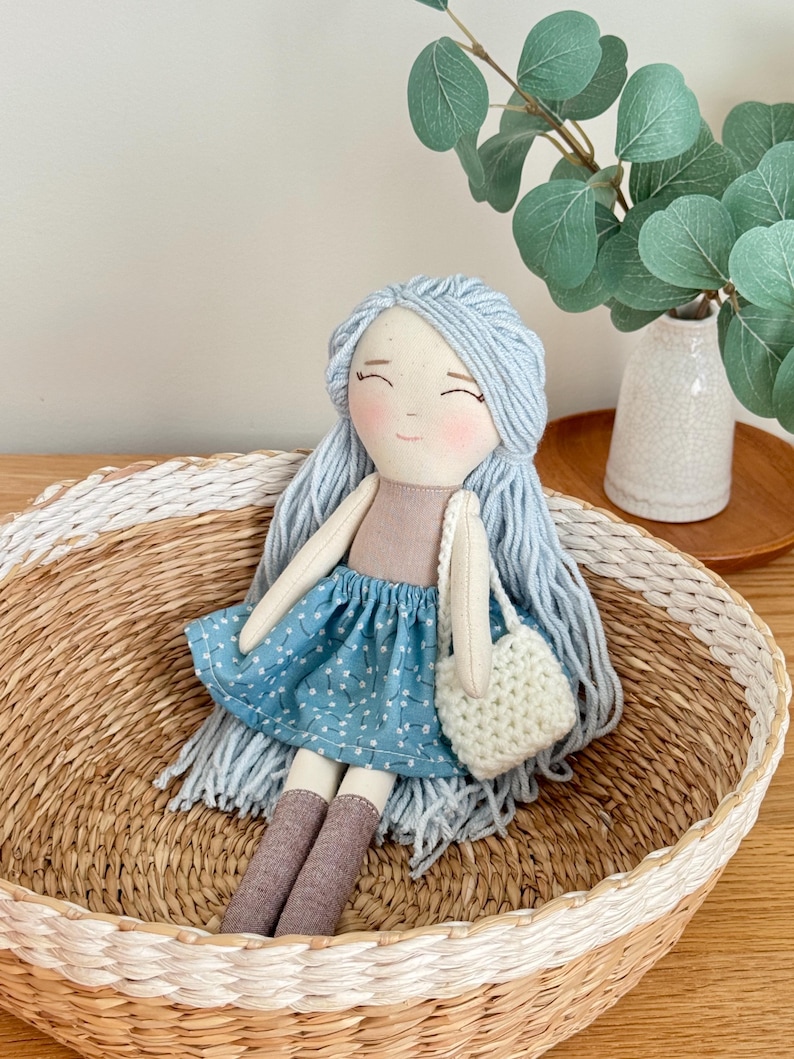 May include: A handmade doll with light blue yarn hair, rosy cheeks, and a blue patterned skirt. The doll is holding a small, white crocheted bag and is seated in a woven basket with a white painted rim. The doll's socks are brown.