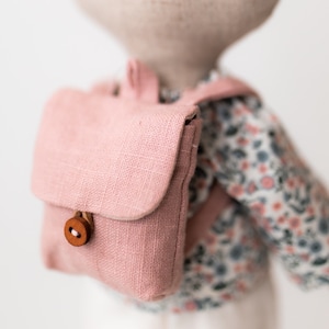 May include: A pink linen backpack with a wooden button closure. The backpack is worn by a doll with a floral print shirt.
