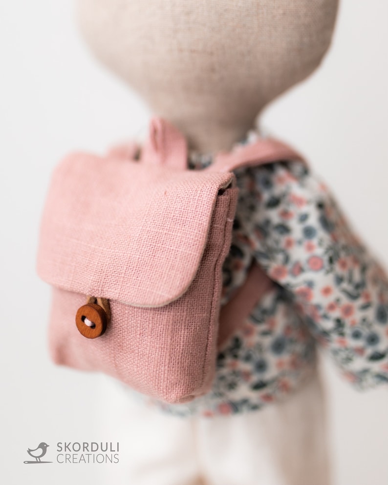 May include: A pink linen backpack with a wooden button closure. The backpack is worn by a doll with a floral print shirt.