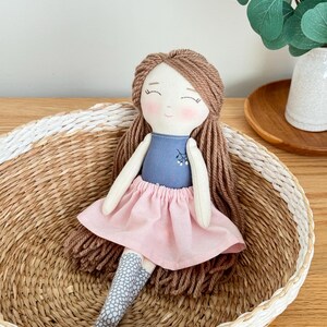 Peut inclure: Poup&eacute;e en tissu faite &agrave; la main avec des cheveux en fil marron, v&ecirc;tue d'un haut bleu et d'une jupe rose. La poup&eacute;e a des joues roses et est assise dans un panier en osier. Les chaussettes de la poup&eacute;e ont un motif gris et blanc.
