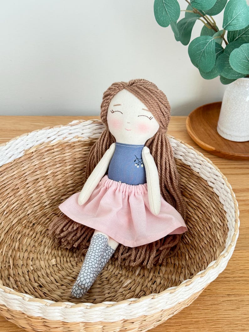 Peut inclure: Poup&eacute;e en tissu faite &agrave; la main avec des cheveux en fil marron, v&ecirc;tue d'un haut bleu et d'une jupe rose. La poup&eacute;e a des joues roses et est assise dans un panier en osier. Les chaussettes de la poup&eacute;e ont un motif gris et blanc.
