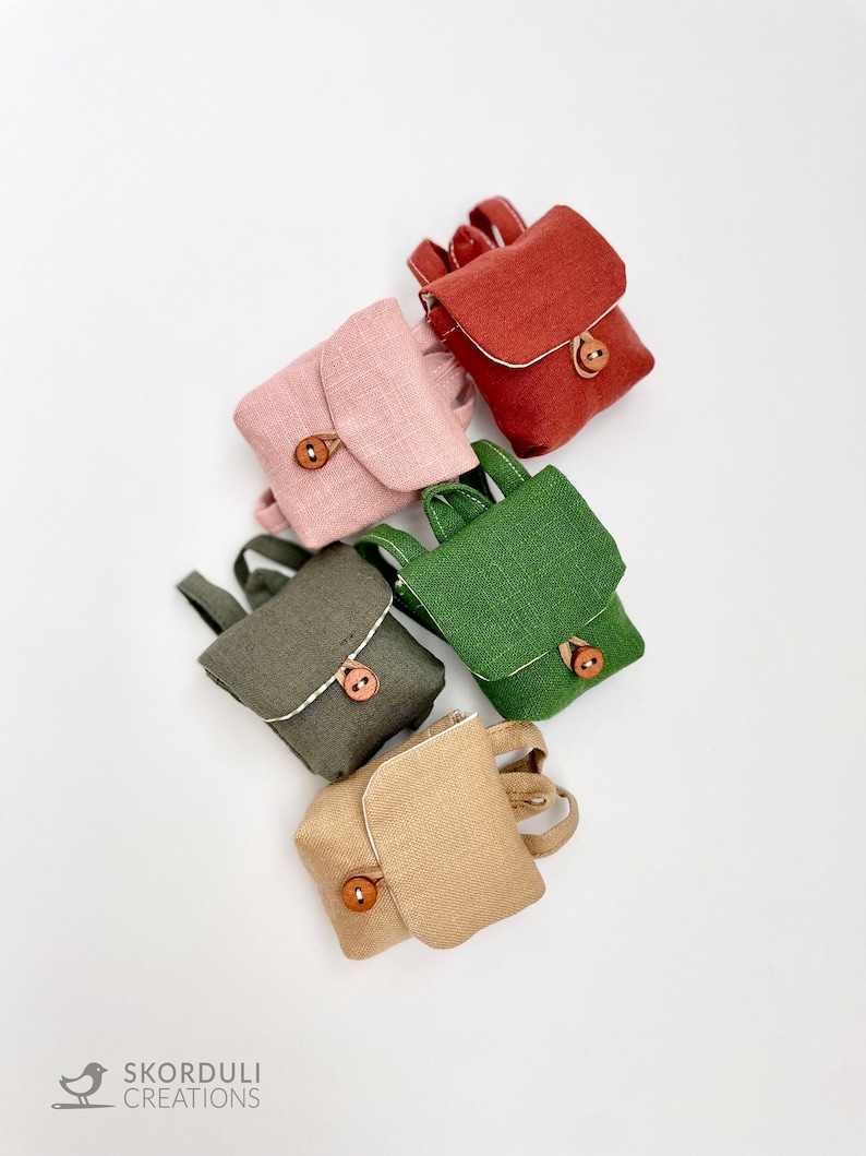 May include: Five miniature linen backpacks in different colors: red, pink, green, brown, and beige. Each backpack has a small wooden button closure.