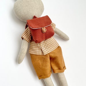 May include: A plush bunny toy wearing a brown and white checkered shirt, brown shorts, and a small brown backpack with a wooden button.