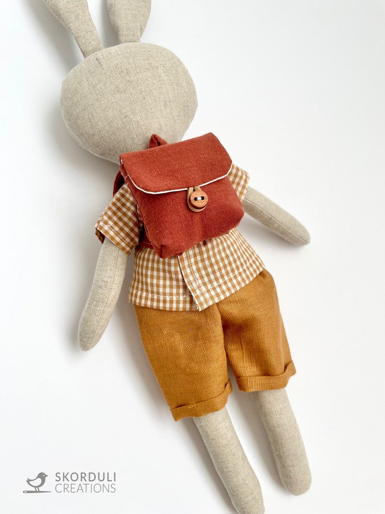 May include: A plush bunny toy wearing a brown and white checkered shirt, brown shorts, and a small brown backpack with a wooden button.