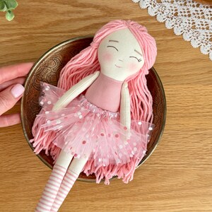 Puede incluir: Una mu&ntilde;eca hecha a mano con cabello de hilo rosa, un vestido rosa y una falda de tul con lunares blancos. La mu&ntilde;eca est&aacute; en un cuenco dorado. La mu&ntilde;eca tiene calcetines a rayas y una cara dulce y sonriente. Un juguete encantador y caprichoso.
