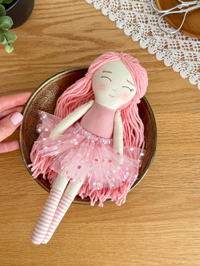 Puede incluir: Una mu&ntilde;eca hecha a mano con cabello de hilo rosa, un vestido rosa y una falda de tul con lunares blancos. La mu&ntilde;eca est&aacute; en un cuenco dorado. La mu&ntilde;eca tiene calcetines a rayas y una cara dulce y sonriente. Un juguete encantador y caprichoso.