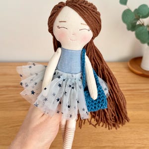 May include: A handmade doll with brown yarn hair and a blue denim top. The doll wears a white tulle skirt with navy blue star patterns and carries a small blue crocheted bag. The doll's face has embroidered features and rosy cheeks.