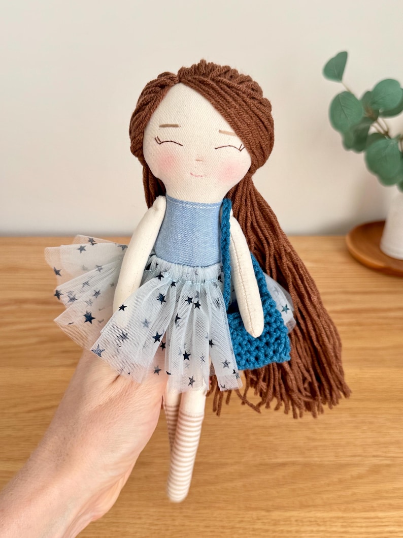 May include: A handmade doll with brown yarn hair and a blue denim top. The doll wears a white tulle skirt with navy blue star patterns and carries a small blue crocheted bag. The doll's face has embroidered features and rosy cheeks.
