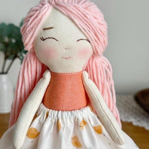 May include: A handmade fabric doll with pink yarn hair and a smiling face. The doll wears an orange top and a white skirt patterned with yellow birds. The doll's arms are white and the overall design is soft and whimsical.