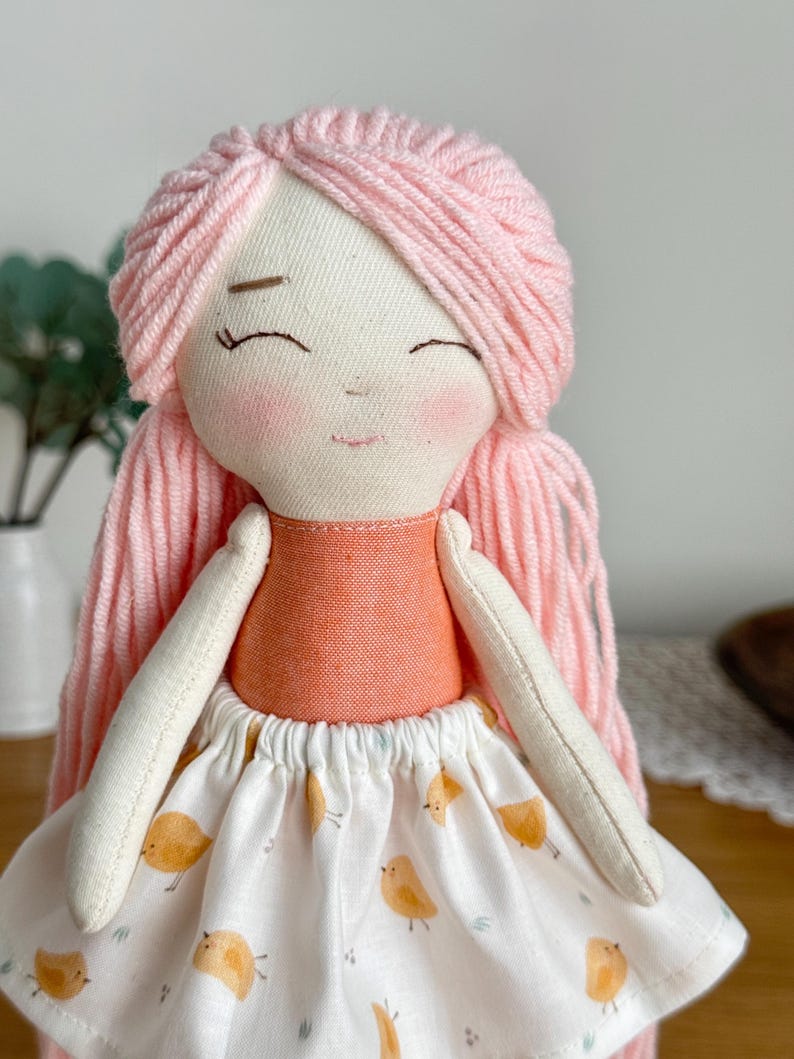 May include: A handmade fabric doll with pink yarn hair and a smiling face. The doll wears an orange top and a white skirt patterned with yellow birds. The doll's arms are white and the overall design is soft and whimsical.