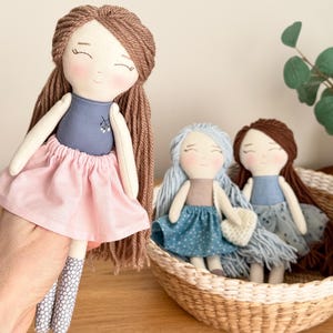 Peut inclure: Poup&eacute;es en tissu faites &agrave; la main avec des visages souriants. Une poup&eacute;e est tenue, v&ecirc;tue d'un haut bleu et d'une jupe rose. Deux autres poup&eacute;es sont assises dans un panier en osier, l'une avec des cheveux et une robe bleus, l'autre avec des cheveux bruns et une robe bleue.