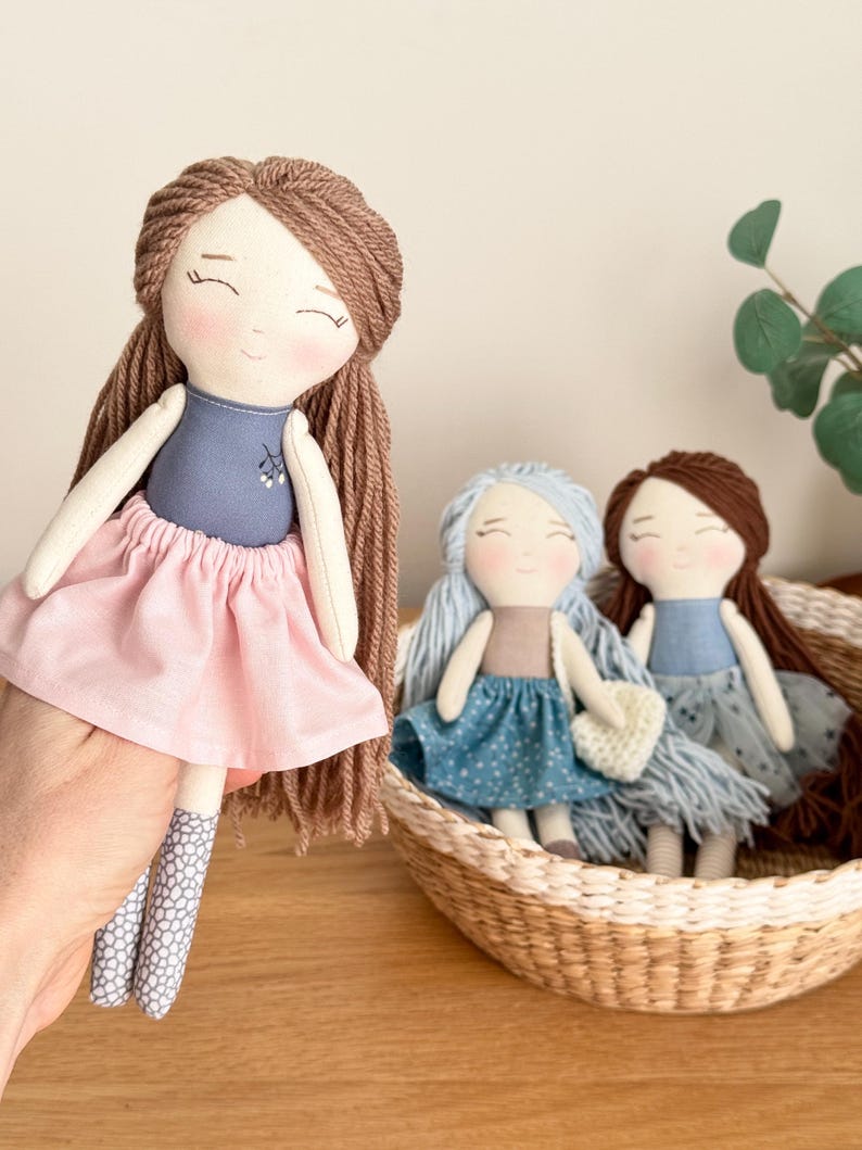 Peut inclure: Poup&eacute;es en tissu faites &agrave; la main avec des visages souriants. Une poup&eacute;e est tenue, v&ecirc;tue d'un haut bleu et d'une jupe rose. Deux autres poup&eacute;es sont assises dans un panier en osier, l'une avec des cheveux et une robe bleus, l'autre avec des cheveux bruns et une robe bleue.