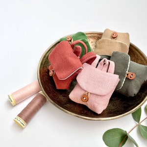 May include: Five miniature fabric bags in various colors, including pink, green, brown, and red, are arranged in a brass bowl. The bags have a flap closure and a small button. Two spools of thread are visible on the left side of the image.