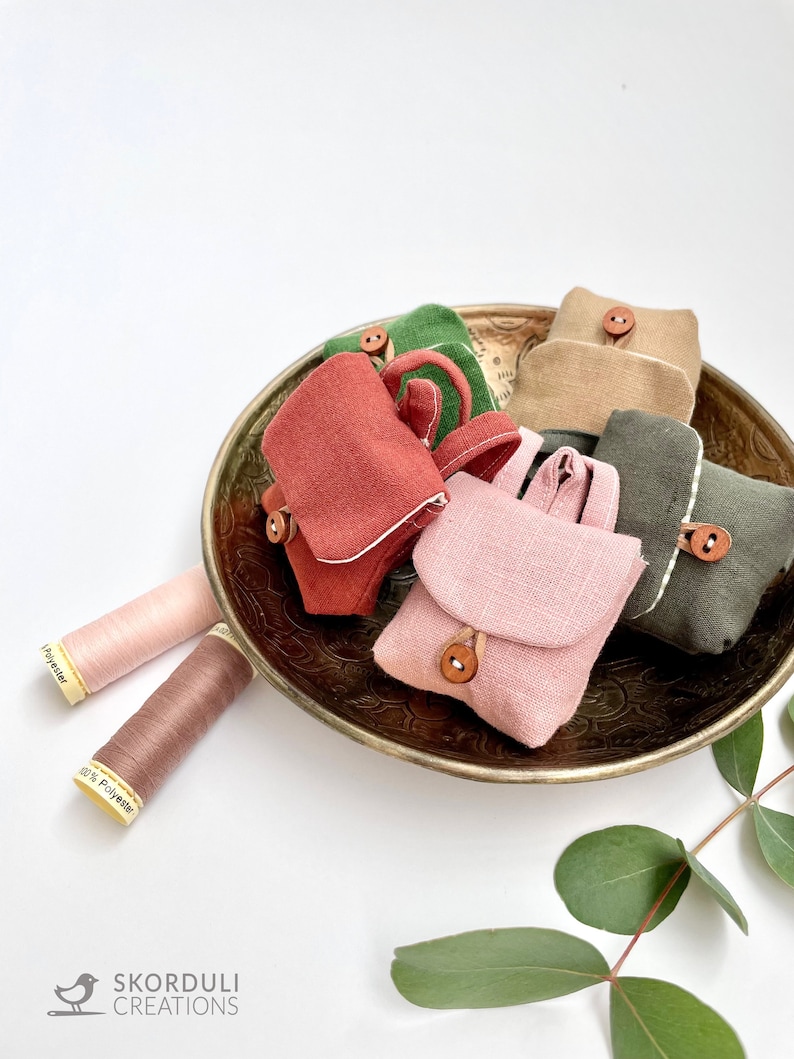 May include: Five miniature fabric bags in various colors, including pink, green, brown, and red, are arranged in a brass bowl. The bags have a flap closure and a small button. Two spools of thread are visible on the left side of the image.
